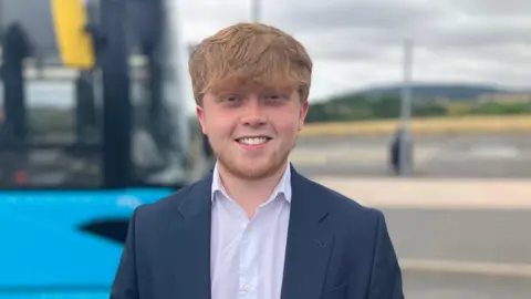Ginger-haired man with a ginger beard, wearing a navy suit and white shirt, smiling in front of a blue single-decker bus, with a car park and hills in the background 