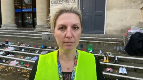 Jo Roberts in a high vis jacket with her blonde hair pulled back and looking at the camera. Behind her are children's wellies and outdoor shoes in pairs with a council building in the background. 