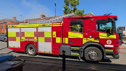 A side view of a fire engine. The red, yellow and silver vehicle is stationary on a road with a red brick building in the background. The scene is set against a blue sky and sunshine.