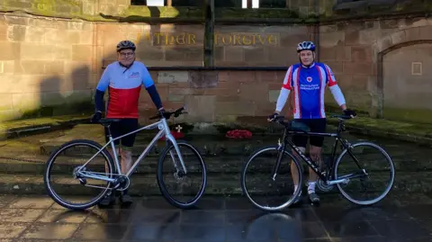 Two men in cycling gear and helmets stand in front of a war memorial holding their road bikes in front of them. Stuart Green on the left is wearing a white and red top, and Roger Churchill on the right is wearing a blue, white and red cycling top.
