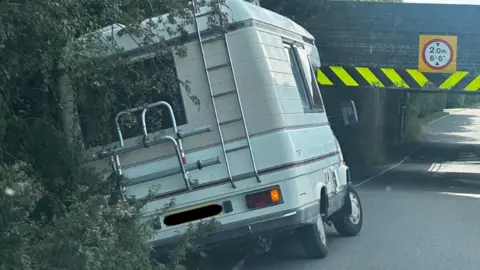 James Fuller A white and beige camper van in a verge at the approach to the 2m (6ft 6inch) high underpass.
A low 2m (6 ft 6 inch) high underpass, with circular red and white warning signs that has unseen railway track above.
The is a yellow and black chevron that is damaged by vehicles hitting.