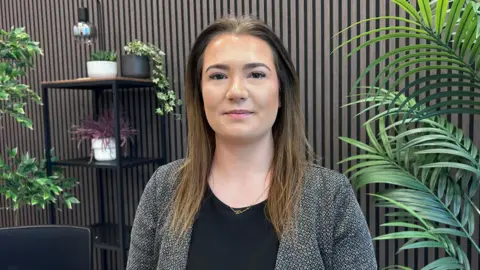 Kate Bradbrook/BBC Det Ch Insp Harrison smiles at the camera standing in a smart office with large green plants and a wood-panelled wall. She wears a black top under an open mottled grey jacket and has long brown hair.