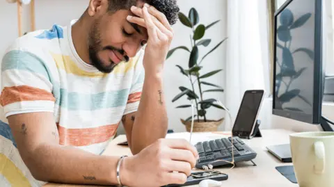 A young man wearing a colourful striped t-shirt rests his head in his hand while sitting at a desk in front of a computer.