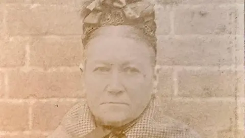 A sepia close-up photograph of a stern faced heavily built woman in her fifties with her hair parted in the middle and mostly hidden by a hat made out of lace with bows on the top. She is wearing a checked shawl and standing in front of a brick wall.