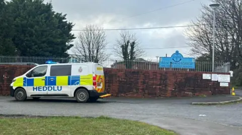 A brown school wall with a silver railing above and a police van parked outside. In the background is a blue school sign.