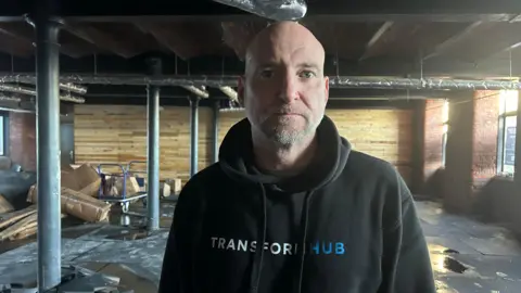 A man with a bald head and beard wearing a black jumper stands in a building which has flooding damage, the floor has wet puddles and boxes and crates are scattered in the left hand corner.