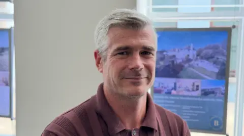 A man with short grey hair wearing a burgundy collared knit shirt, seated indoors near a window. Behind them, blurred property advertising displays show photographs of homes and buildings. Natural light enters through the window, creating a bright office setting.