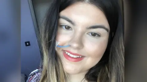 Ayla Jones A head and shoulders shot of Ayla, who is white, has long brown hair, lightened at the ends, with brown eyes. She is looking slightly below the line of the camera but is smiling slightly towards it. She has a blue naso-gastric tube coming out of her right nostril which is taped to her cheek.