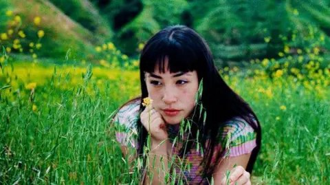 Ana Chen A picture of Liang sat in a field of short grass/flowers. She is wearing a pink outfit, she has long black hair.