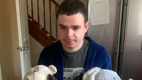 Jo Brearton A young man aged in his 20s and with short, brown cropped hair sits clutching several cuddly toys. He wears a blue dressing gown.