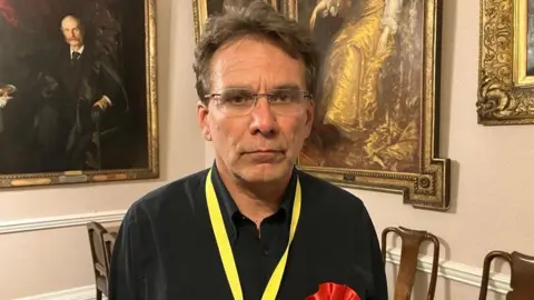 A head and shoulders shot of Darren Hale looking glum at the camera. He is wearing a black shirt, a yellow lanyard around his neck and a red badge. He has short brown hair and is also wearing glasses.