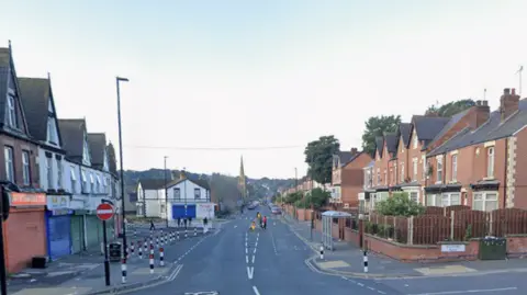 An empty suburban street with small shops with shutters down to the left and terraced homes to the right.