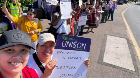 A group of UNISON members stand along a roadside holding flags and placards during a union demonstration. Several signs call for fair pay, respect for workers, and the restoration of overtime arrangements. Participants are gathered in a long line, with purple UNISON branding visible throughout the scene. The event takes place on a sunny day, with supporters displaying banners and signs to highlight workplace concerns.