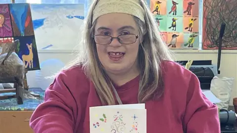 Julia Gregory/BBC A young woman dressed in pink is holding a Christmas card she's made