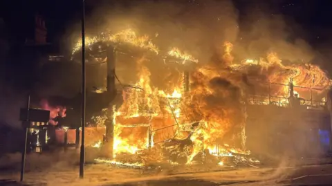 Essex Fire and Rescue Service A fire is earing through a two storey building and has burnt down the walls, just leaving the bones and foundations of the site. It remains engulfed in flames. The sky is dark making the fire appear even brighter against the night sky.