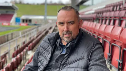 James Burridge/BBC Kelvin Thomas, in a black quilted jacket, seated in an empty football stadium. 