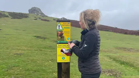 Dartmoor National Park Association Ella Breins stood to the right of a post with two signs on it. She is looking at the sign and is wearing a black jumper, gilet and trousers. Behind the post is the moors. 