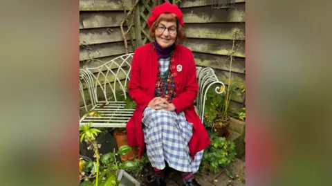 Contributed Marcia Riddington wears a red hat with a long red cardigan. She has red hair and black glasses and is sitting on a garden bench. Beneath her cardigan she is wearing a chequered blue and white dress with a multi-coloured vest over it. 