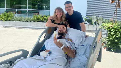 Luke Tarrant Luke Tarrant, lying in a hospital bed, outside, eating an ice cream, with a bandaged arm, tubes coming out his other arm, with his parents, Daniela and David Tarrant behind him and behind a bed. David has his arm around Daniela, he is wearing a dark T-shirt and Daniela, a floral dress, with long fair hair. David is wearing glasses. 