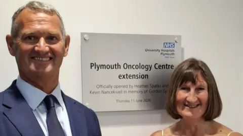 A smartly dressed man and woman standing side-by-side smile toward the camera. They are standing in front of a plaque which says Plymouth Oncology Centre extension officials opened by Heather Sparks and Kevin Nancekivell in memory of Gordon Sparks.
