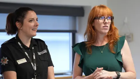 A woman with dark hair tied in a ponytail is wearing a black t-shirt and lanyard. her t-shirt says "fire control". next to her in angela rayner, who has bright ginger hair and is wearing a green dress