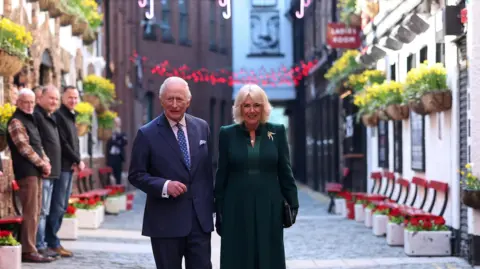 Reuters King Charles and Queen Camilla during a visit to Commercial Court, Belfast. The street has a cobbled ground, the buildings either side have red benches outside and flower baskets hang outside. A group of people are to the left watching the King and Queen walk down the street. The king has white hair, wearing a blue pin stripe suit with light blue pocket square, white shirt and blue polka dot tie. The queen has blonde hair, wearing a green coat gold bird brooch, black gloves, and carrying a black clutch. 