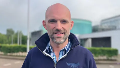 A man who is bald with a black and grey beard looks into the camera. He is smiling and is wearing a blue check shirt with a navy pullover. The pullover has the emblem of a fox on the front.