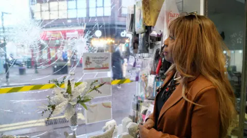 Diane stands inside her shop, facing a large front window that’s been shattered and is marked with yellow caution tape. Shelves of beauty products and a glittery silver floral decoration sit nearby. Through the cracked glass, a busy street with pedestrians and colourful storefronts is visible.