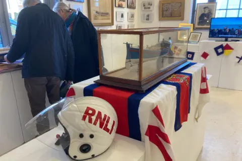 Luke Deal/BBC An old white RNLI helmet next to a display case with a model of an old lifeboat inside 