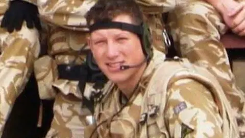 Family handout A young man in the army who smiles at the camera. He wears camouflage uniform and wears a head strap with a microphone attached to it. 