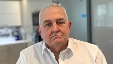 Brown has short grey hair and is sitting in his kitchen wearing a white shirt.