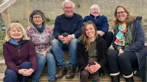 A group of people are all smiling at the camera. They are sitting on some steps in a walled garden. They are all wearing warm clothing and boots for gardening. 