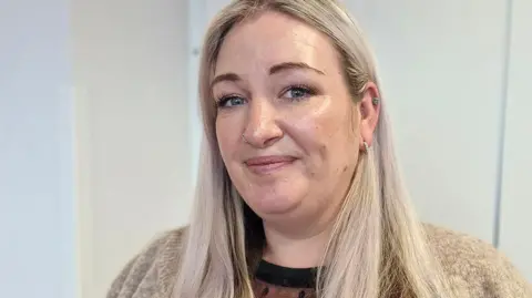 Hayley Winstanley with long blond hair wearing a sand colour cardigan and black top underneath. She has a nose ring and two earrings. She is smiling.