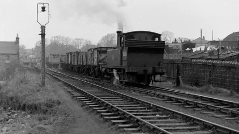 Don Martin Black and white picture showing road traffic waiting at Whitegates Level Crossing while a steam locomotive crosses in 1960