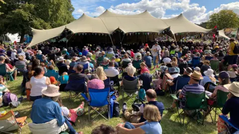Martin Heath/BBC Crowds sit outside a large open sided marquee facing towards it, listening to a speaker. Many wear straw hats and sit in camping chairs. There are trees around and the sky is blue with white clouds.