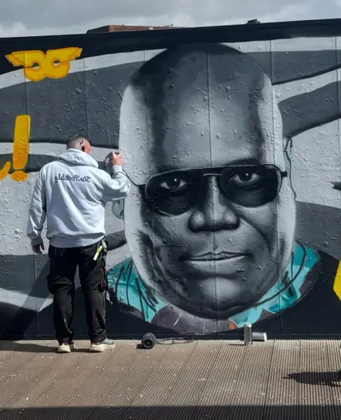 Steve Jenkins Jenkins is a white man with short dark hair and is seen from behind working on the mural. He holds a can of spray paint and wears a grey hoodie and black trousers and trainers.