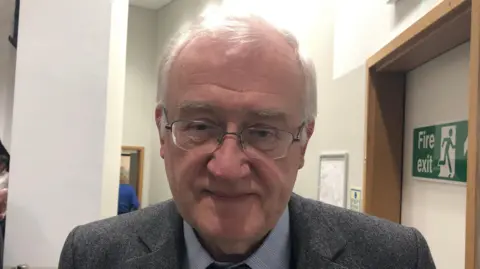 Vikki Irwin/BBC Christopher Hudson smiles at the camera. He has grey hair and glasses. He wears a grey suit with a light blue shirt and blue tie.