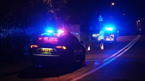 Eddie Mitchell A picture of two police cars at the scene of a stabbing in Pulborough Road, Cootham, near Storrington, on the evening of Thursday, 16 January. A police officer can be seen at the boot of the furthest car forward.