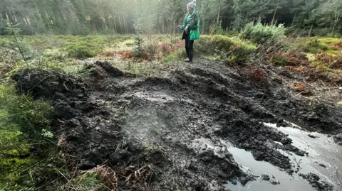 North York Moors National Park Authority A woman in a green coat and hate stands on top of a mound of earth where the landscape has been damaged by an off-road vehicle. 