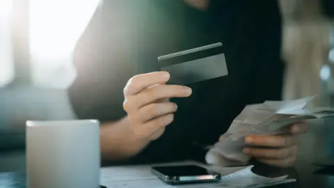 Getty Images Fotografía recortada de una joven asiática sosteniendo una tarjeta de crédito y recibos de gastos, entregando información bancaria y financiera personal en casa.