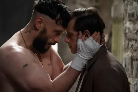 BBC/Mam Tor Productions/Anne Binckebanck A man with a beard and bandages on his hands holds the face of a man wearing a suit.