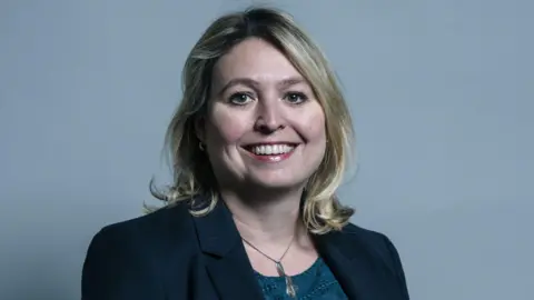 UK Parliament A smiling Dame Karen Bradley has fair shoulder-length hair and is smiling. She is wearing a dark blue suit jacket over a turquoise top and a metallic necklace.