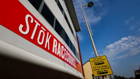 Outside of the STōK Racecourse. A red banner is across the building which reads 'STōK Racecourse' in capital letters. The font is bold and white. A lamp post can be seen next to the building which has a yellow and black coach drop-off sign on it.