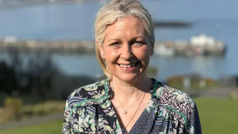 Tiffany Kerruish, who has long blonde hair tied back and is wearing a green, black and white floral top. She is smiling and wearing two necklaces. You can see Douglas Bay in the background behind on a bright sunny day.
