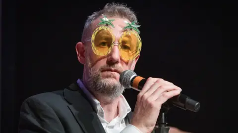 Hollis Photography A man with grey hair is wearing yellow pineapple glasses and has a grey beard. He is holding a microphone and staring into the distance. 