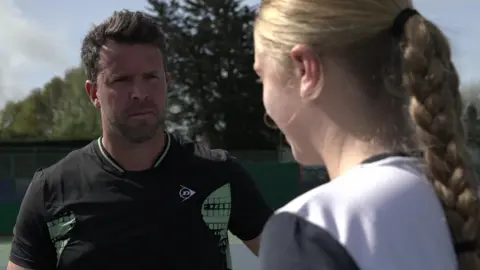 Richard Irwin in a black and green t-shirt stood on a tennis court talking to Daniella who is wearing a blue and white t-shirt.