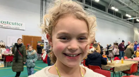 Katie in the community hall, she has tied up curly blonde hair.