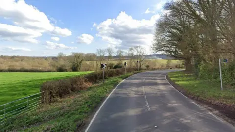 The image shows the turning point of a road with trees on the right-hand-side of the road, while there are bushes on the other side. Plains of grass can also be seen on the left-hand-side. The sky is blue with a few white clouds.