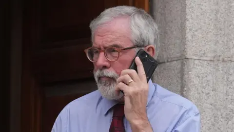 Former Sinn Fein president Gerry Adams holding a mobile phone to his ear outside the High Court in Dublin.  He has short, grey hair and a grey beard.  He is wearing brown-rimmed glasses, a blue shirt and a red patterned tie. 
