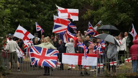 Reuters People hold flags as they gather near a hotel in Woolston as they protest over asylum seekers being housed in the hotel, in Warrington, on 30 August 30, 2025.